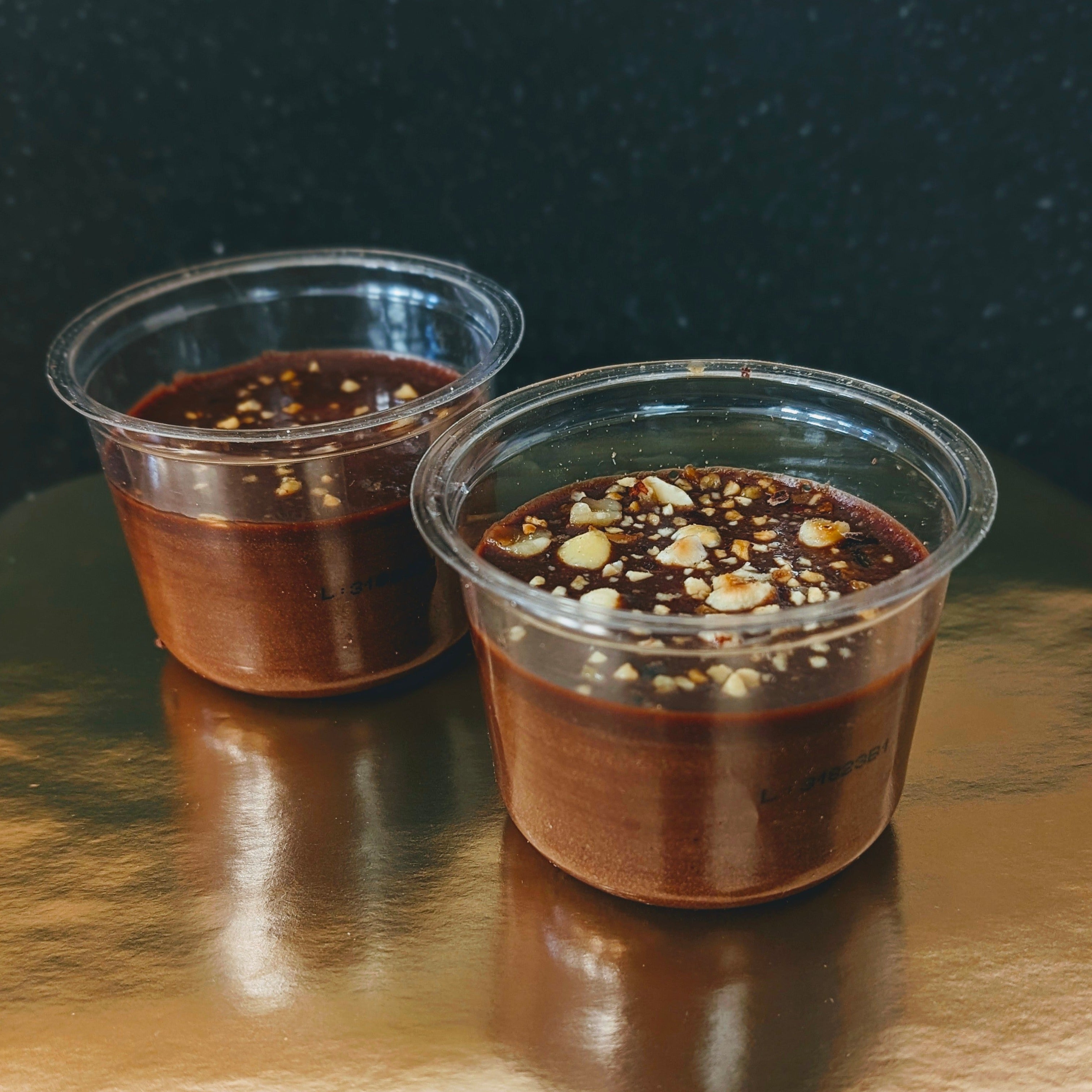 Two clear plastic cups filled with chocolate dessert topped with chopped nuts on a reflective surface.