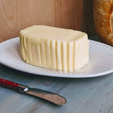 Stick of butter on a white plate next to a butter knife and a loaf of crusty bread on a light wooden table.