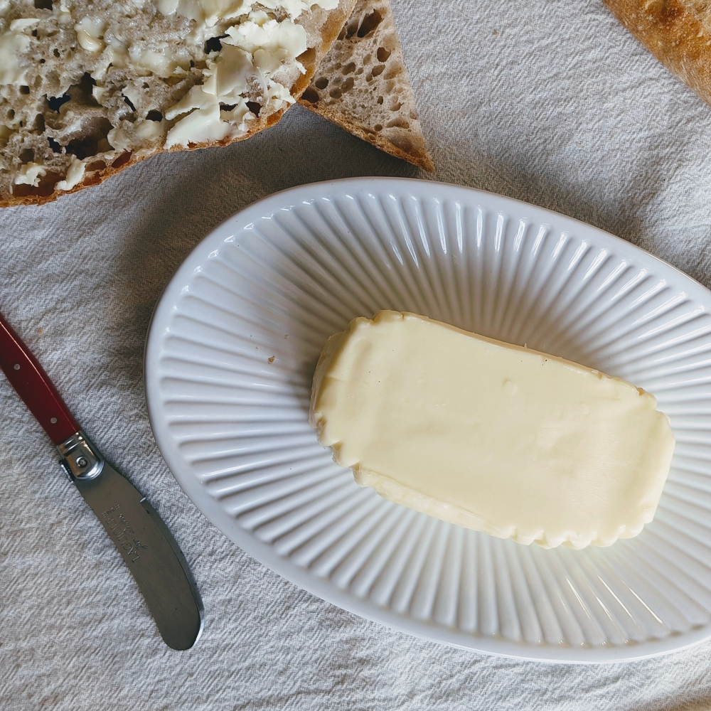 Rectangular stick of butter on a white ridged dish, torn buttered sourdough and a red-handled butter knife on linen.