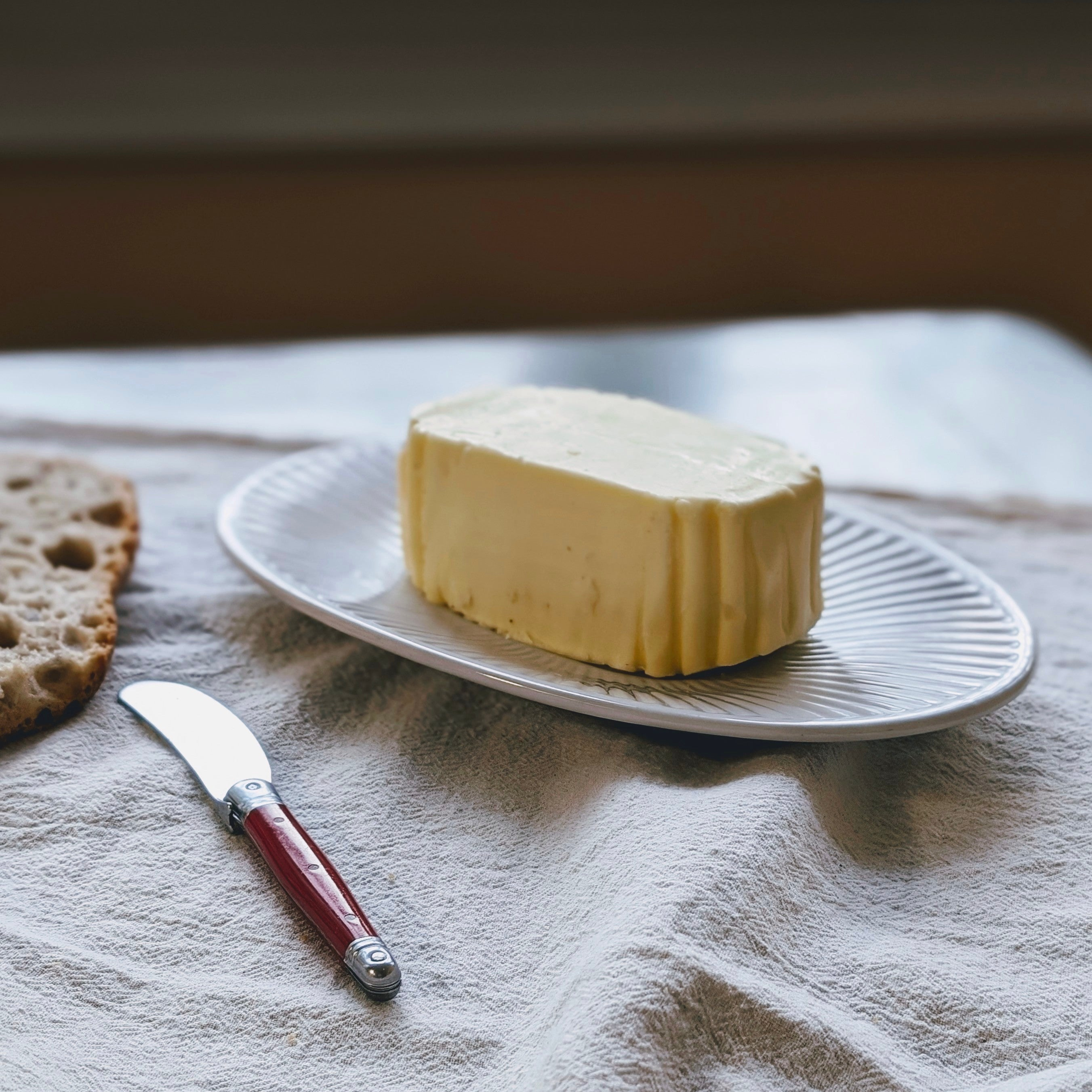 Oval plate with a block of butter, folded cloth beneath, a red-handled butter knife and slice of bread at left.
