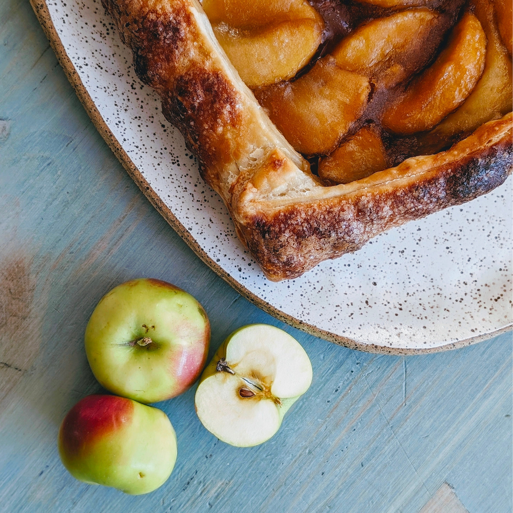 Rustic apple tart on a speckled plate with three fresh apples (one halved) on a blue wooden table.