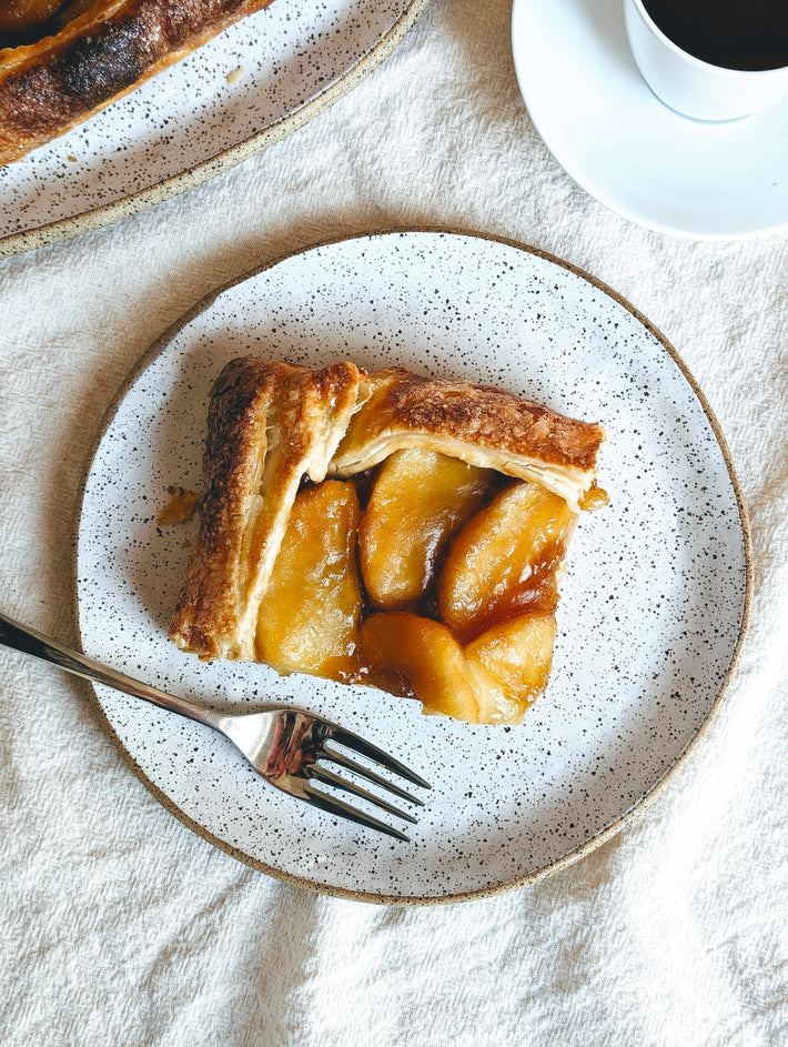 Slice of apple tart with caramelized apple slices on a speckled plate, fork beside it and a coffee cup at top-right.