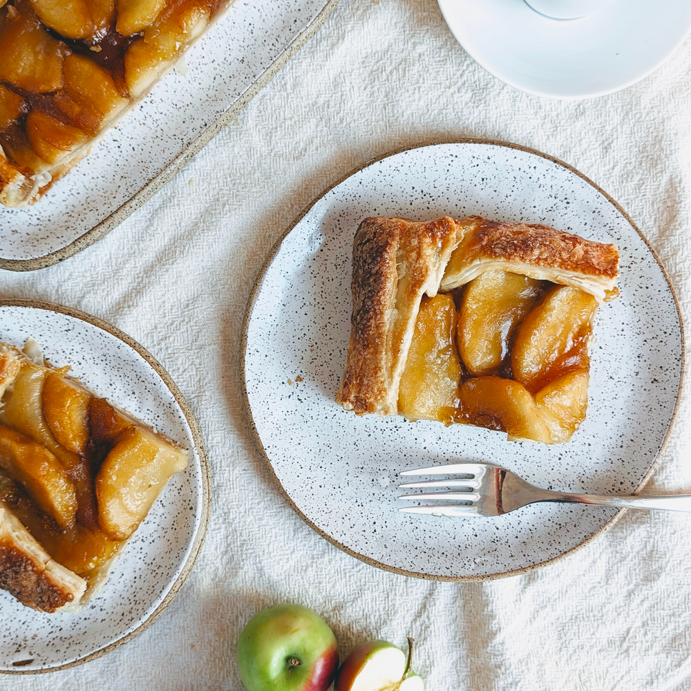 Caramelized apple tart slice on speckled plate with fork; extra tart slices and two apples on cloth.