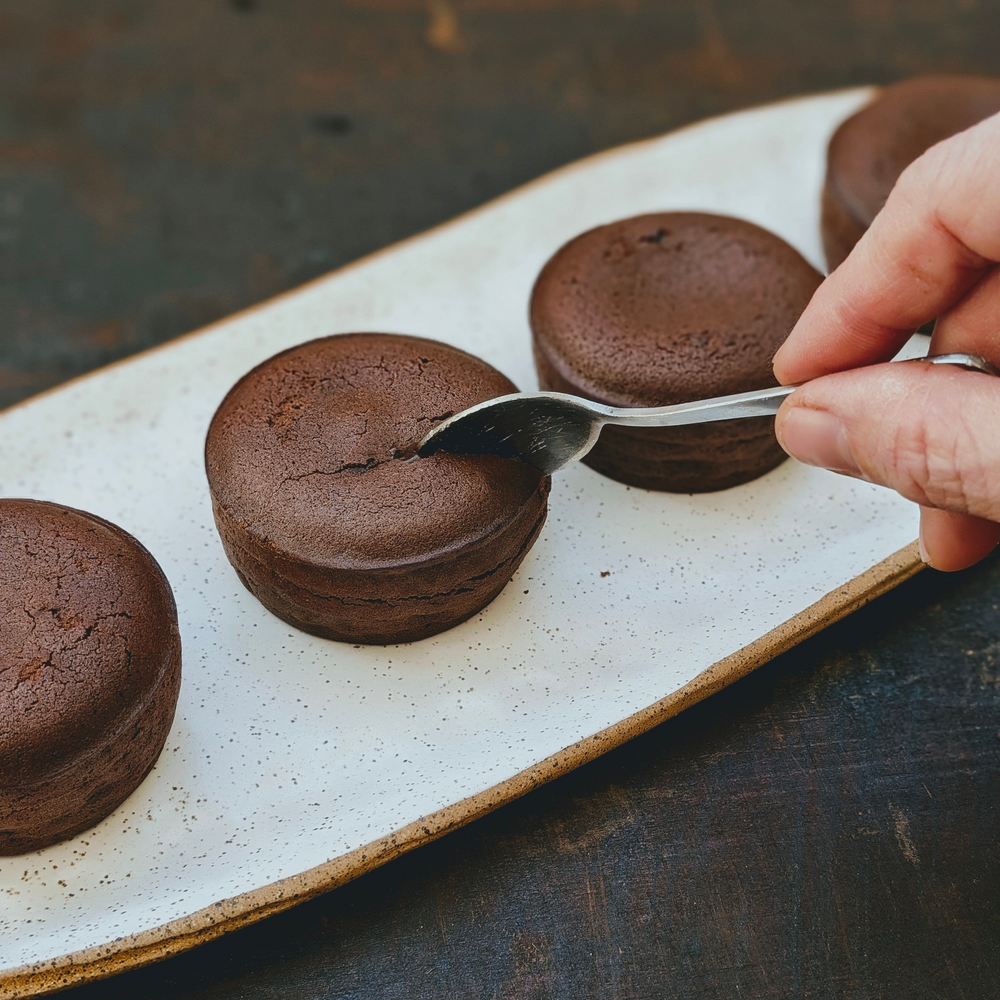 Four round chocolate cakes on a speckled plate; a hand uses a spoon to scoop the center of one.