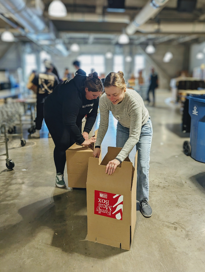 Two volunteers packing cardboard boxes labeled 'Family Meal Box' in a warehouse sorting area