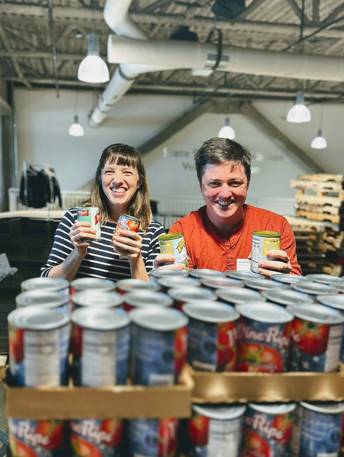 Two smiling volunteers hold canned goods behind stacked cans labeled 'Vine Ripe' in a warehouse food pantry.