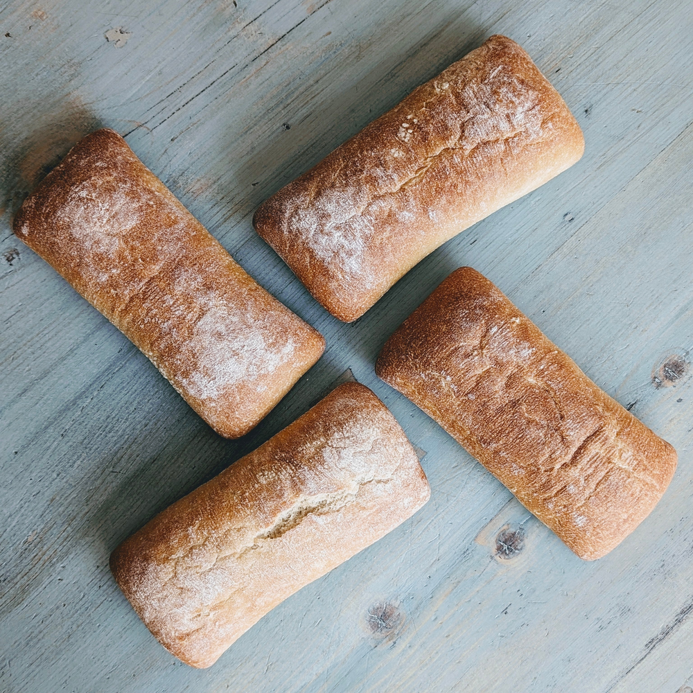 Four dusted ciabatta rolls arranged in a loose square on a weathered blue-gray wooden table.