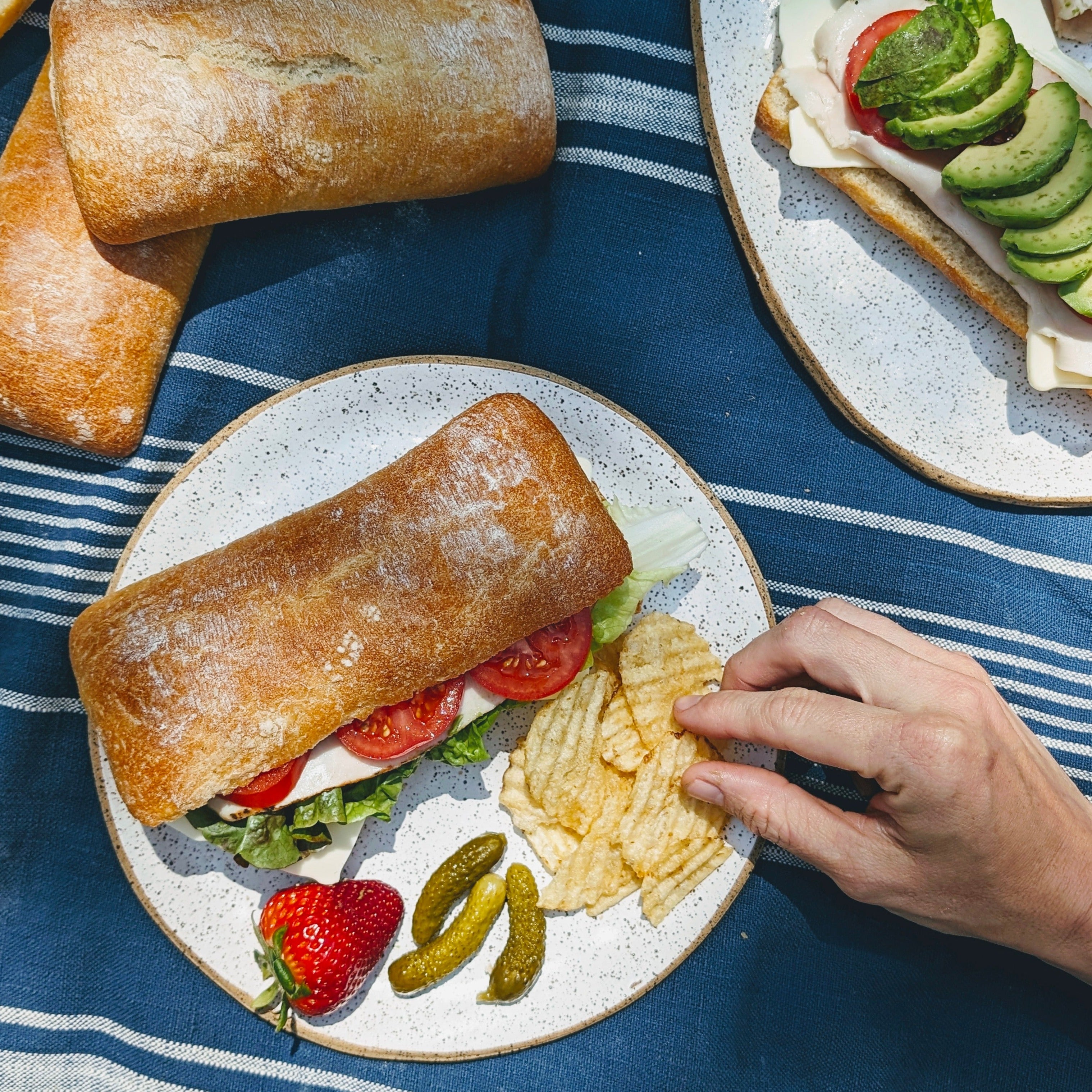 Ciabatta sandwich with lettuce, tomato, cheese on plate with potato chips, cornichons, strawberry; hand reaching for chips.