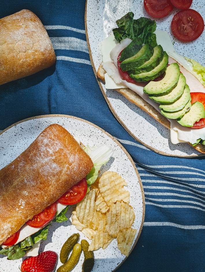 Two ciabatta sandwiches with lettuce, tomatoes, avocado, and chips on a blue tablecloth.
