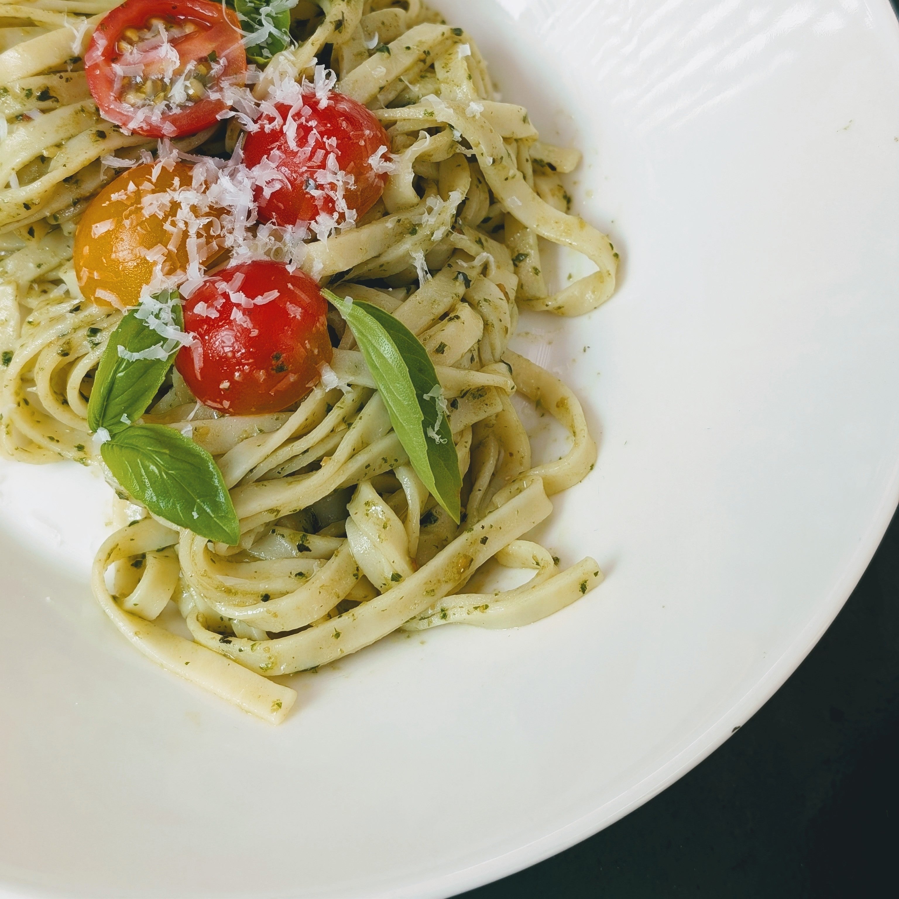 Plated gluten-free linguine pasta with pesto sauce, cherry tomatoes, fresh basil leaves, and grated cheese on a white plate.