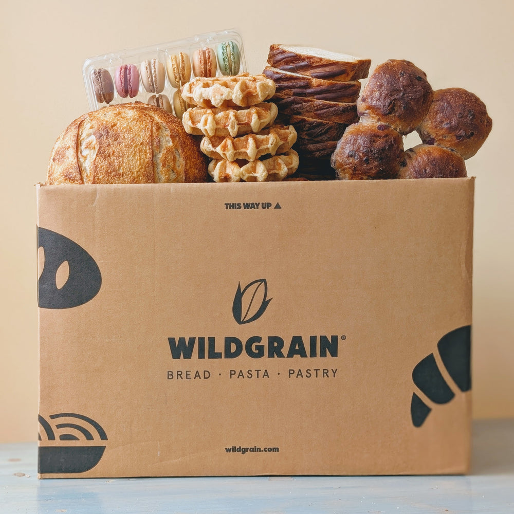 Front view of Wildgrain delivery box showcasing assorted breads, waffles, pastries and macarons with beige background