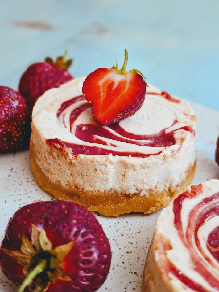Mini strawberry swirl cheesecake on speckled plate with halved strawberry on top and whole strawberries around.