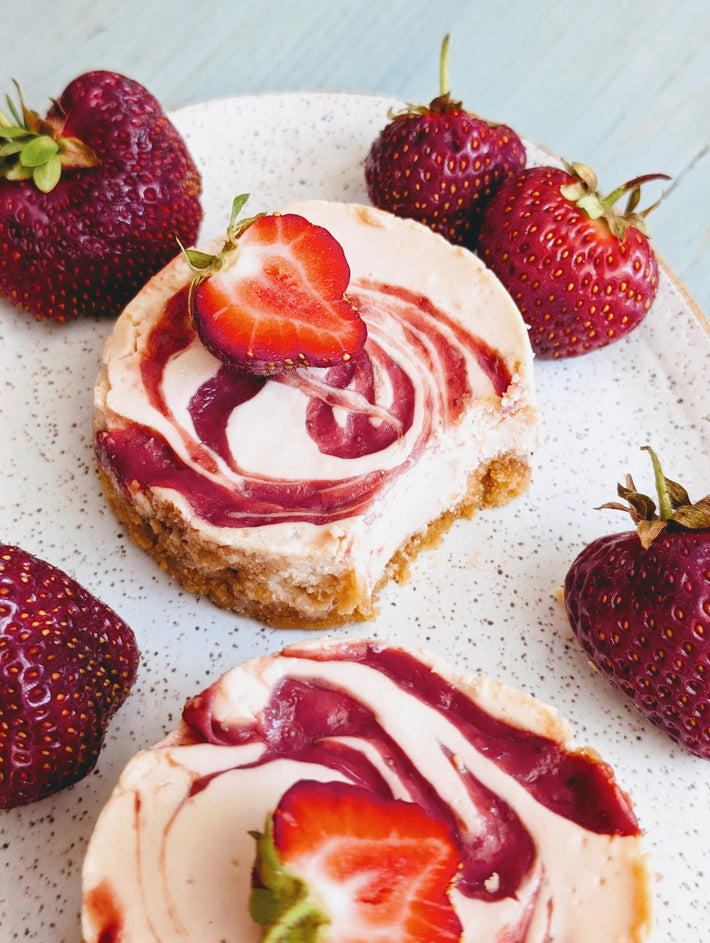 Mini strawberry-swirl cheesecakes on a speckled plate with halved and whole fresh strawberries