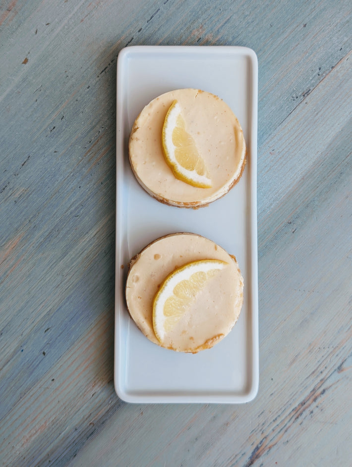 Two lemon-topped cheesecake rounds on a rectangular white plate, top-down view.