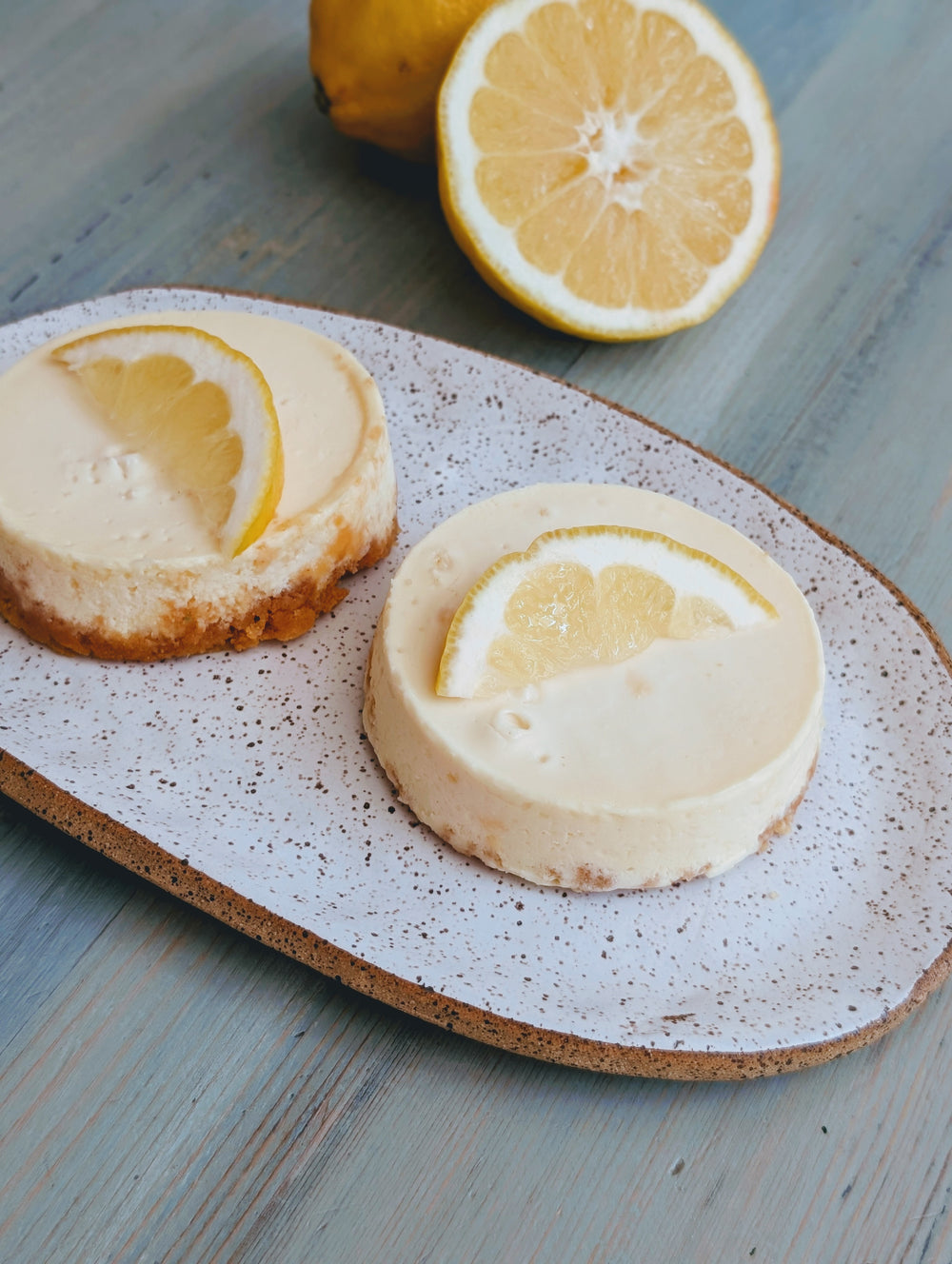 Two mini lemon cheesecakes topped with lemon slices on a speckled oval plate; halved lemon in background.