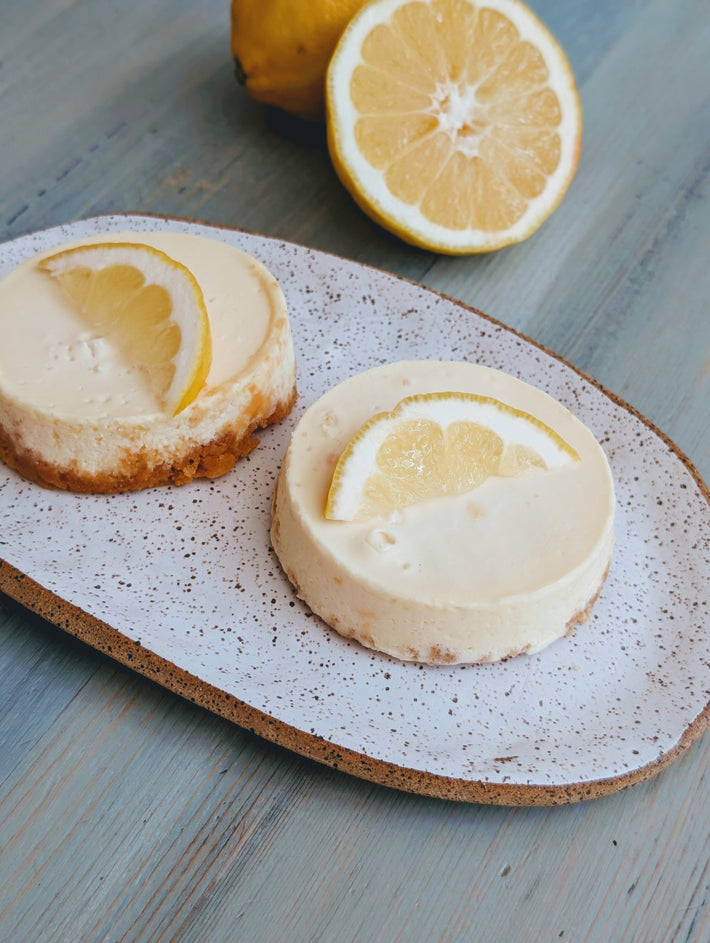 Two mini lemon cheesecakes topped with lemon slices on a speckled oval plate; halved lemon in background.