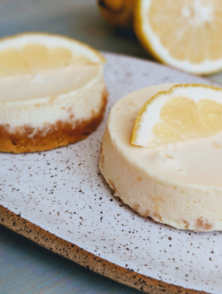 Two mini lemon cheesecakes on a speckled plate, each topped with a lemon wedge.