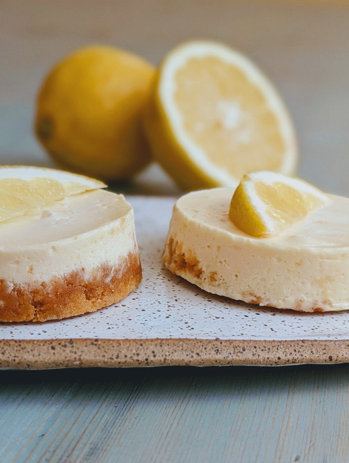 Two mini lemon cheesecakes with graham crust on a speckled plate, lemon slices and whole lemons blurred in background
