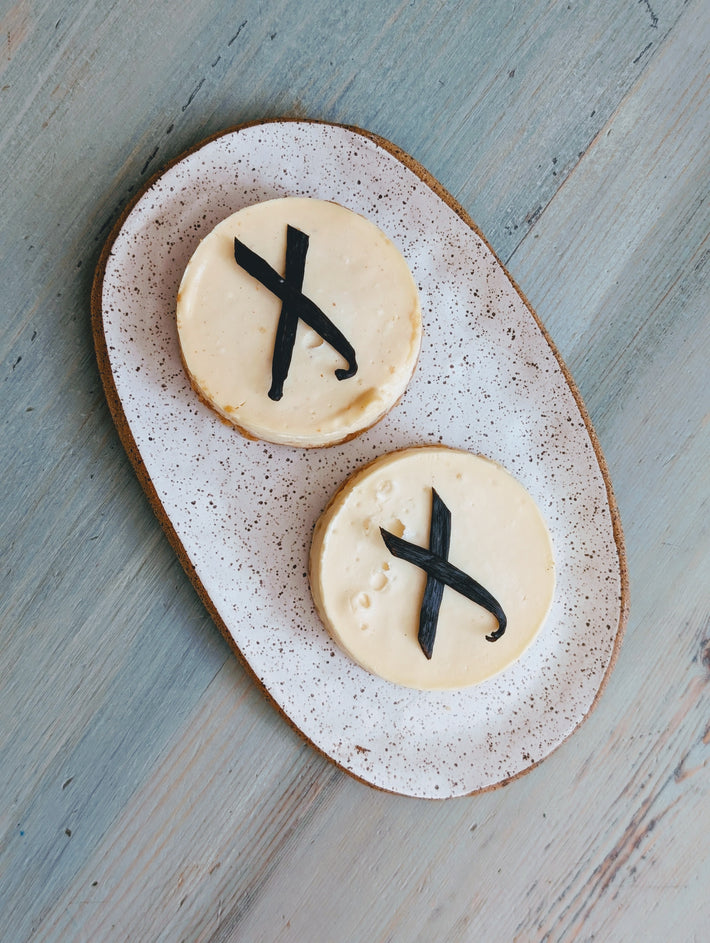 Two round vanilla cheesecakes on an oval speckled plate, each topped with a black vanilla bean arranged like an X.