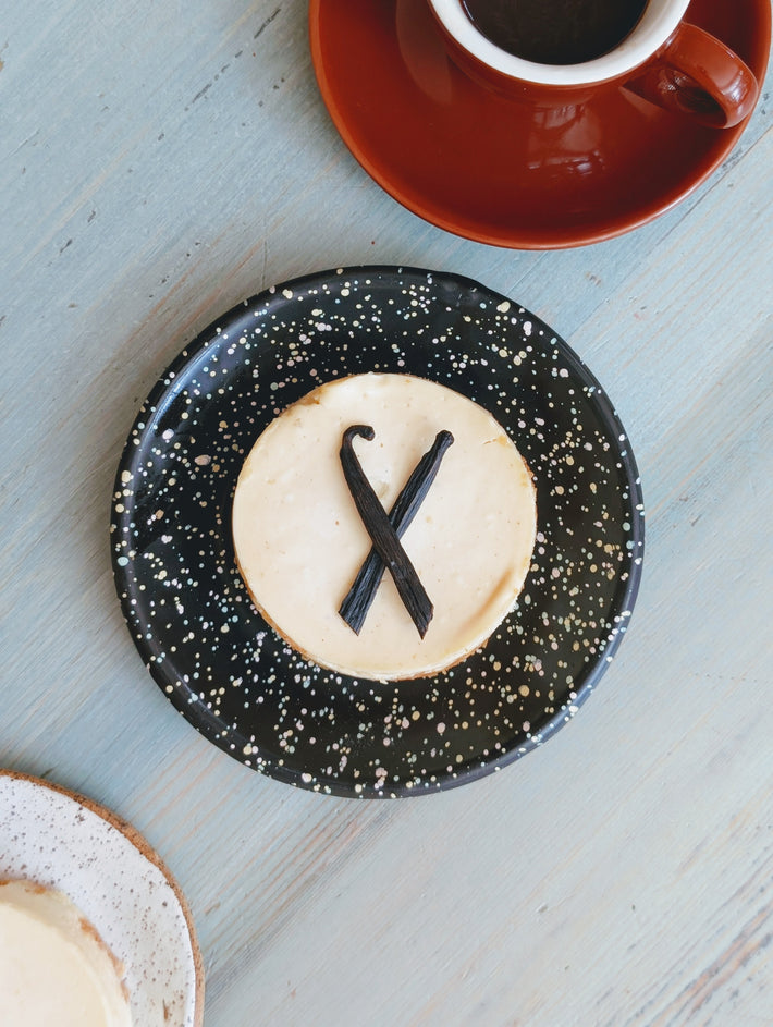 Vanilla cheesecake with two crossed vanilla beans on a speckled black plate; red coffee cup at top right on pale blue table.