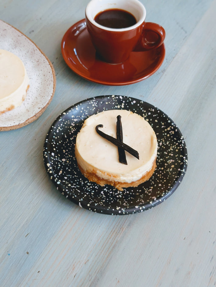 Mini vanilla cheesecake with two vanilla bean pods crossed on a speckled black plate beside an espresso cup.