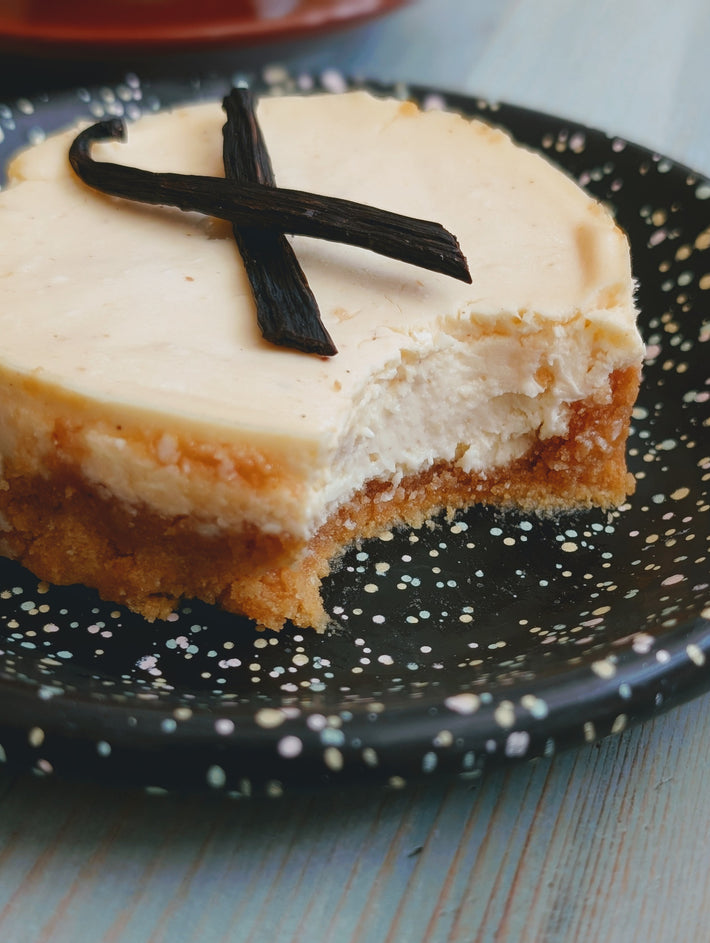 Small slice of vanilla cheesecake with bite taken, topped with two crossed vanilla beans, on a speckled black plate.