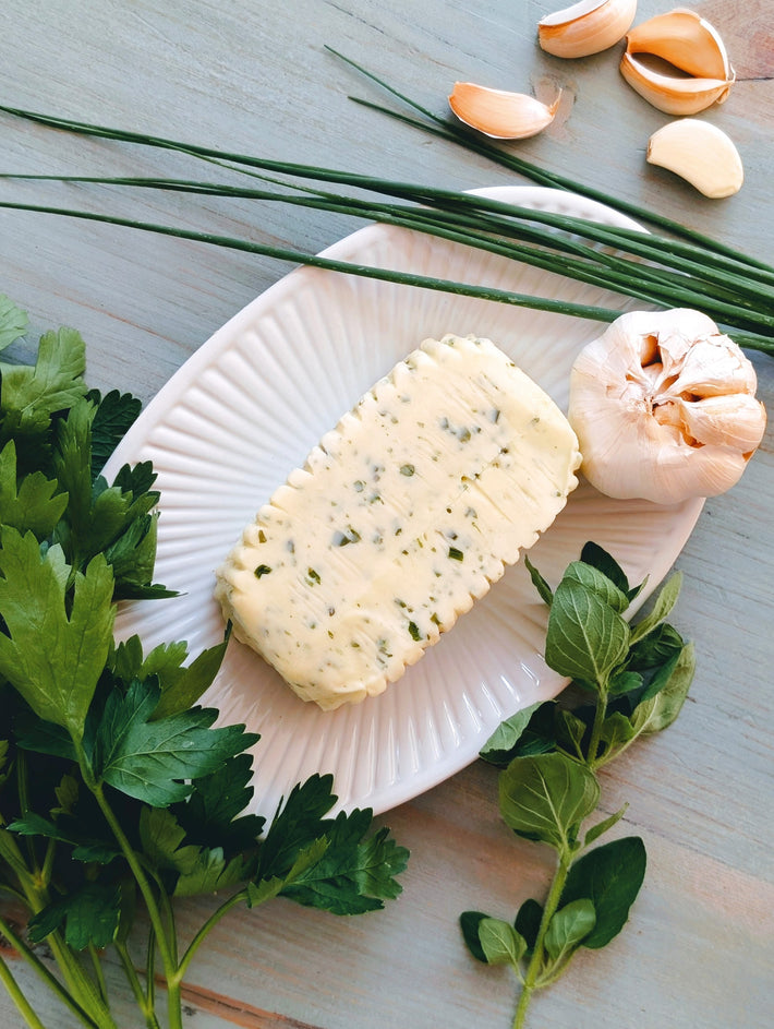 Rectangular herb-speckled butter on white fluted plate, garnished with garlic, chives, parsley and oregano on pale wood.
