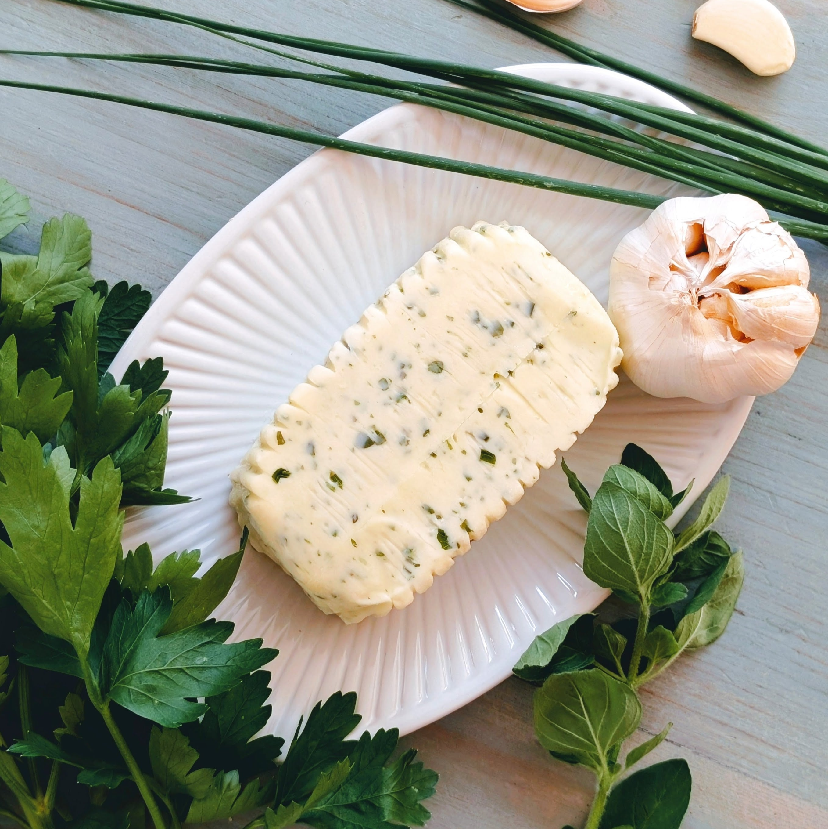 Block of herb butter on a white ridged plate with garlic, chives and fresh parsley and oregano on a wooden surface