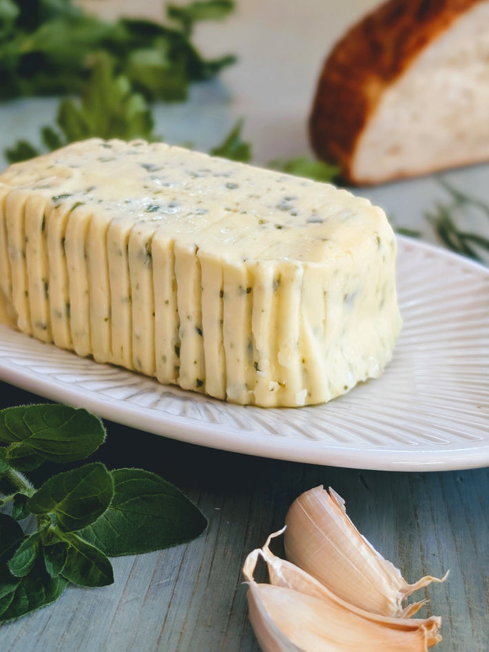 Herb-studded butter log on a white plate, garlic cloves in front and bread with herbs blurred in the background.