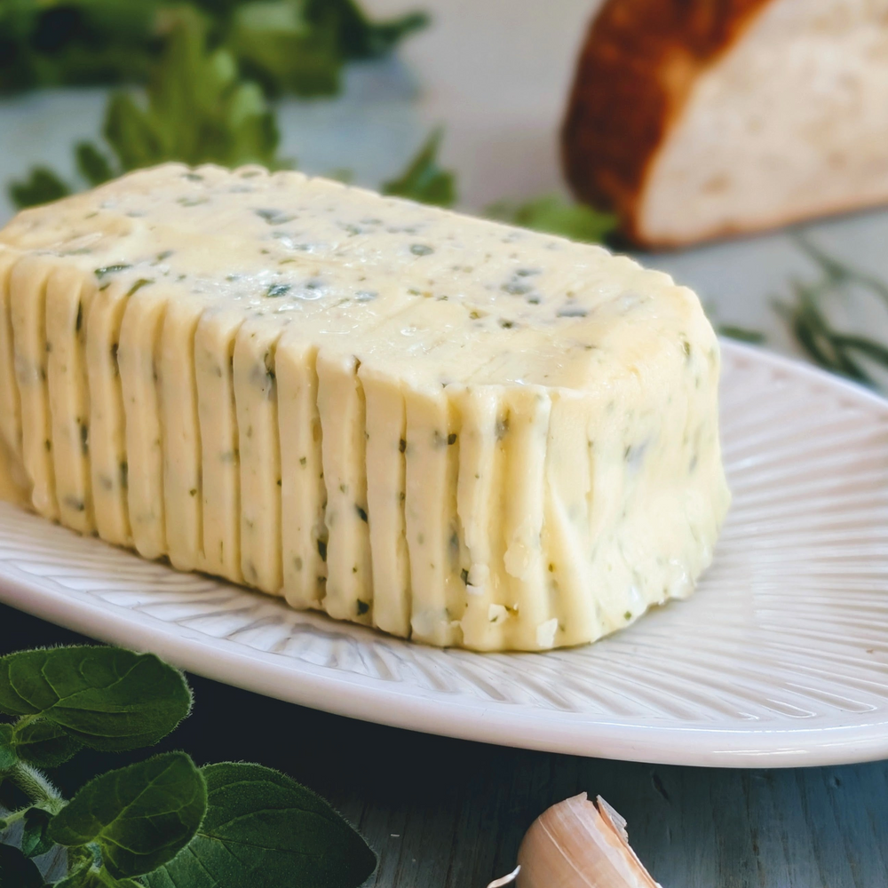 Block of herb butter with visible chive flecks on a white plate, parsley leaves, garlic clove and bread in soft focus