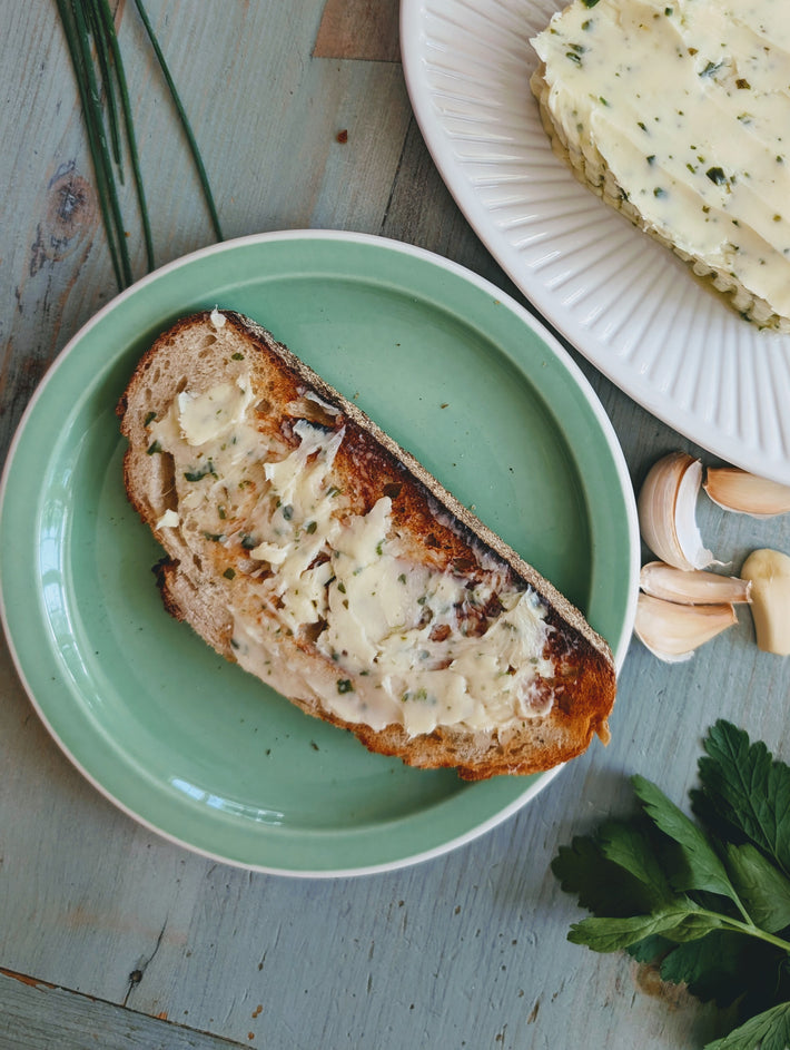 Toasted sourdough slice spread with herbed garlic butter on a green plate, with garlic cloves and parsley beside it.