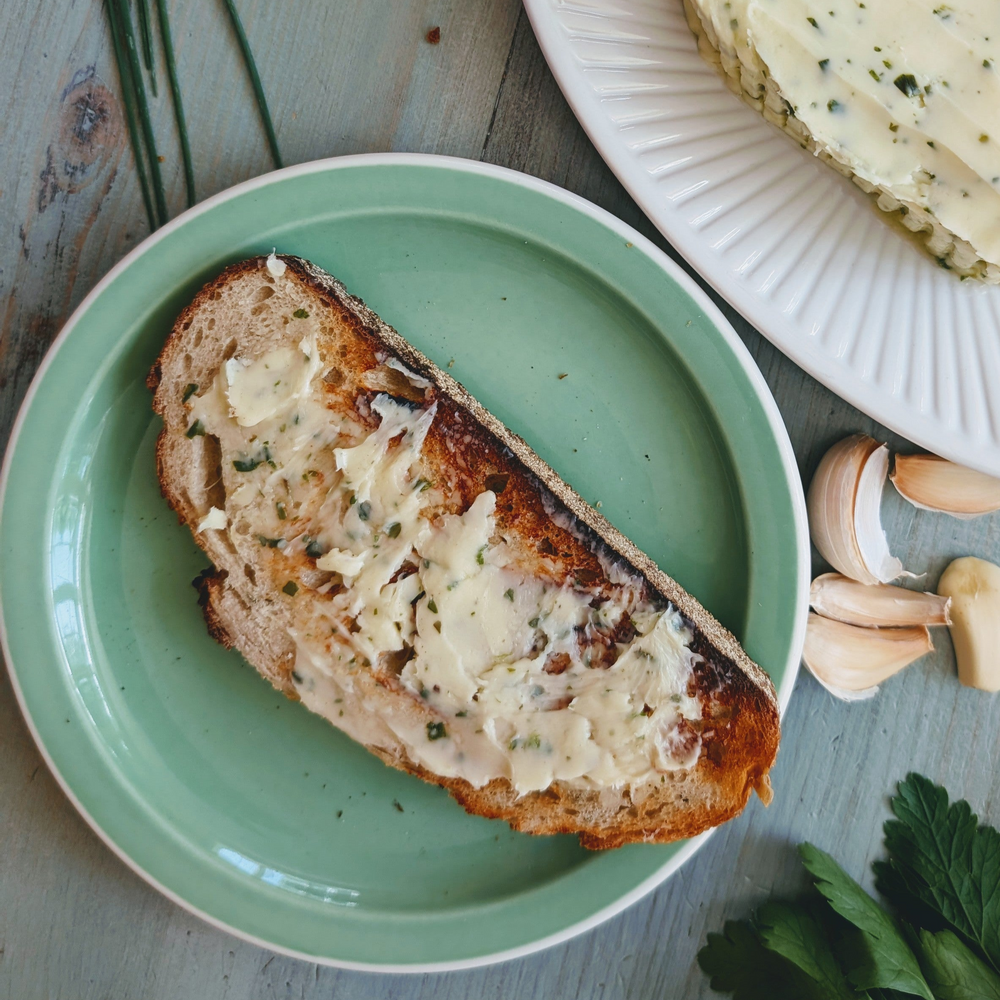Toasted sourdough slice spread with herbed butter on a green plate, garlic cloves and parsley nearby.