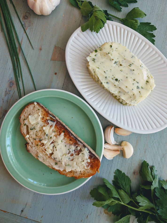 Toasted sourdough slice smeared with garlic-herb butter; nearby block of herb butter on white plate, garlic cloves and parsley.