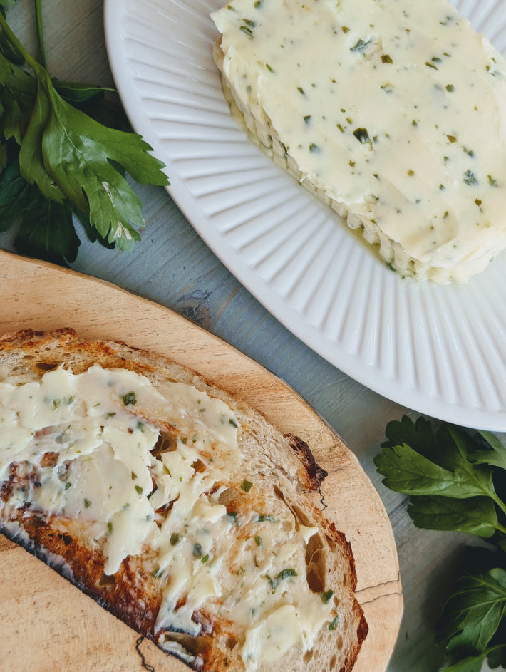 Toasted rustic bread spread with herbed butter, sliced parsley butter on a white plate with parsley sprigs.