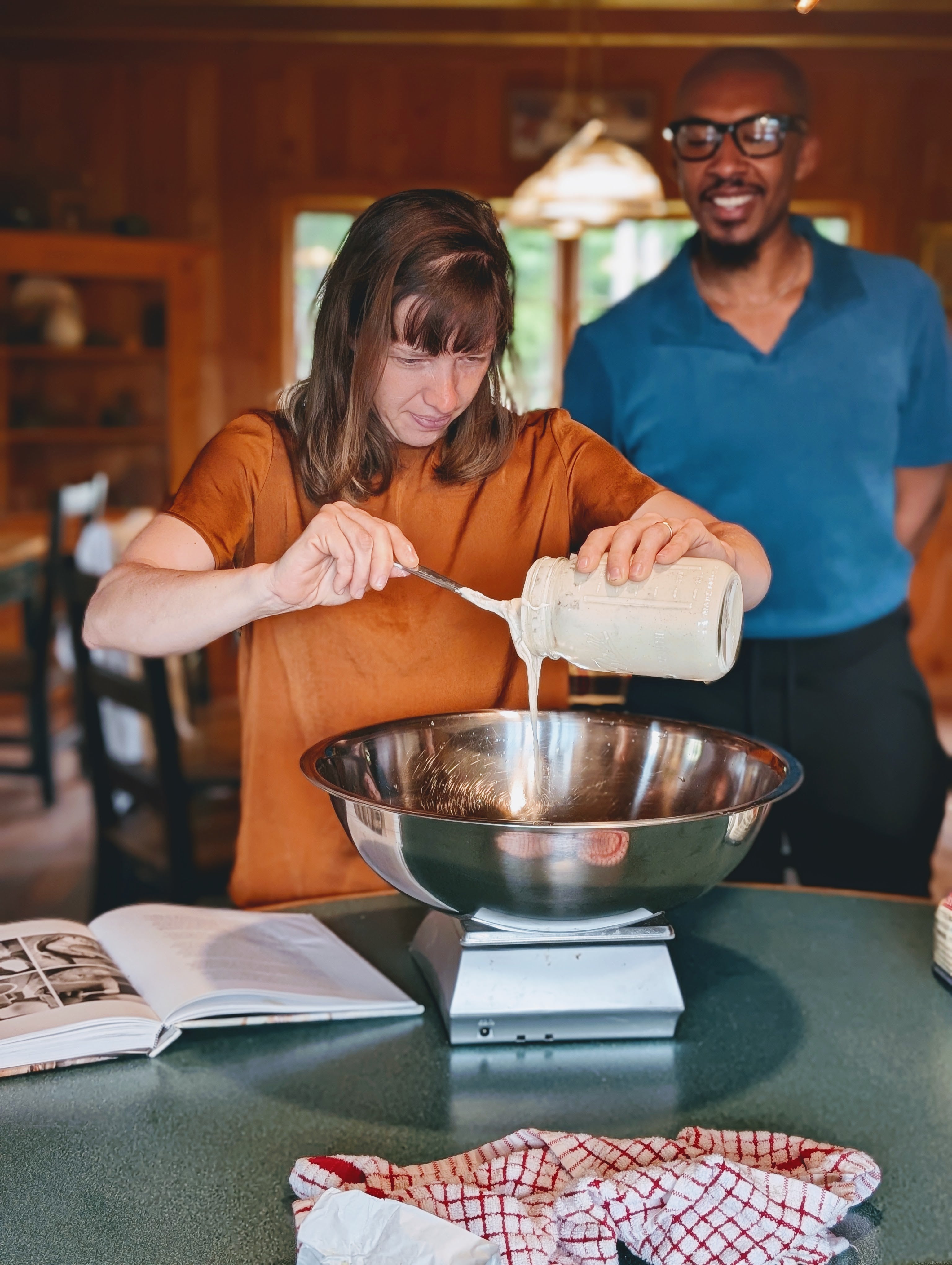 Woman scraping batter from a jar into a metal mixing bowl on a kitchen scale, man smiling behind her