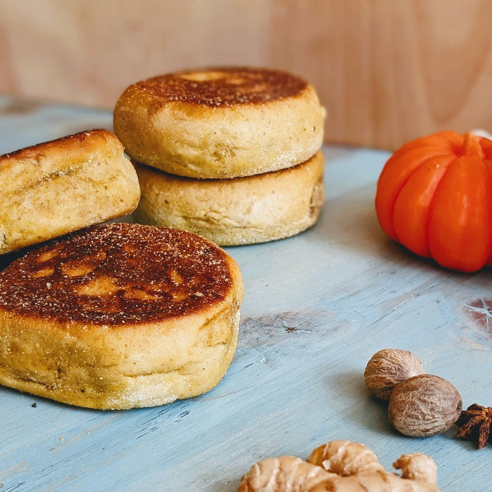 Toasted English muffins stacked on a blue wooden board, with a small decorative pumpkin and whole spices (nutmeg, ginger) nearby.