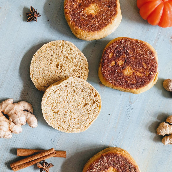 Toasted English muffins, halved and whole, on blue surface with cinnamon sticks, star anise, ginger and a small pumpkin.