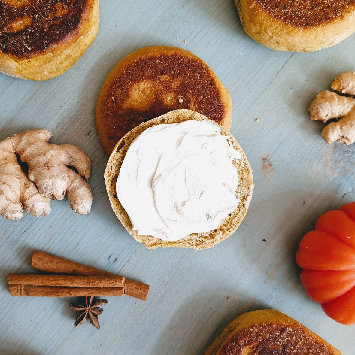 Toasted English muffin halves topped with cream cheese, with ginger, cinnamon sticks, star anise and a small pumpkin on a wood surface.