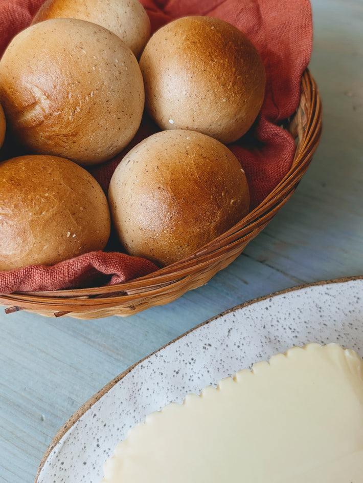 Round whole-wheat dinner rolls in a wicker basket lined with red cloth; speckled plate with a slab of butter at the edge.