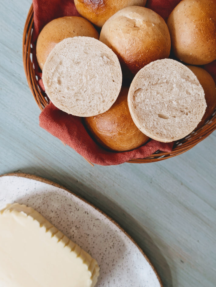 Basket of golden dinner rolls, two cut in half to show crumb, on red linen; speckled ceramic plate with a block of butter