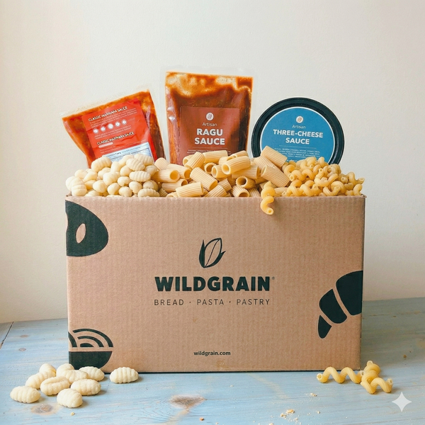 Front view of Wildgrain Pasta 3-Way box with three pasta shapes and three sauce packets on a light wood table