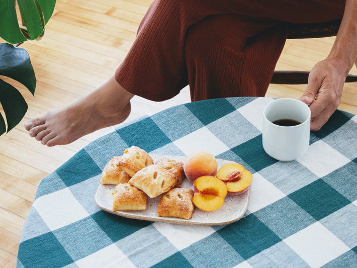 A plate of peach pie bites, fresh peaches, and a cup of coffee on a checkered tablecloth