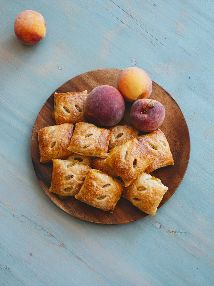 A wooden plate with baked pastries and fresh peaches on a blue surface.