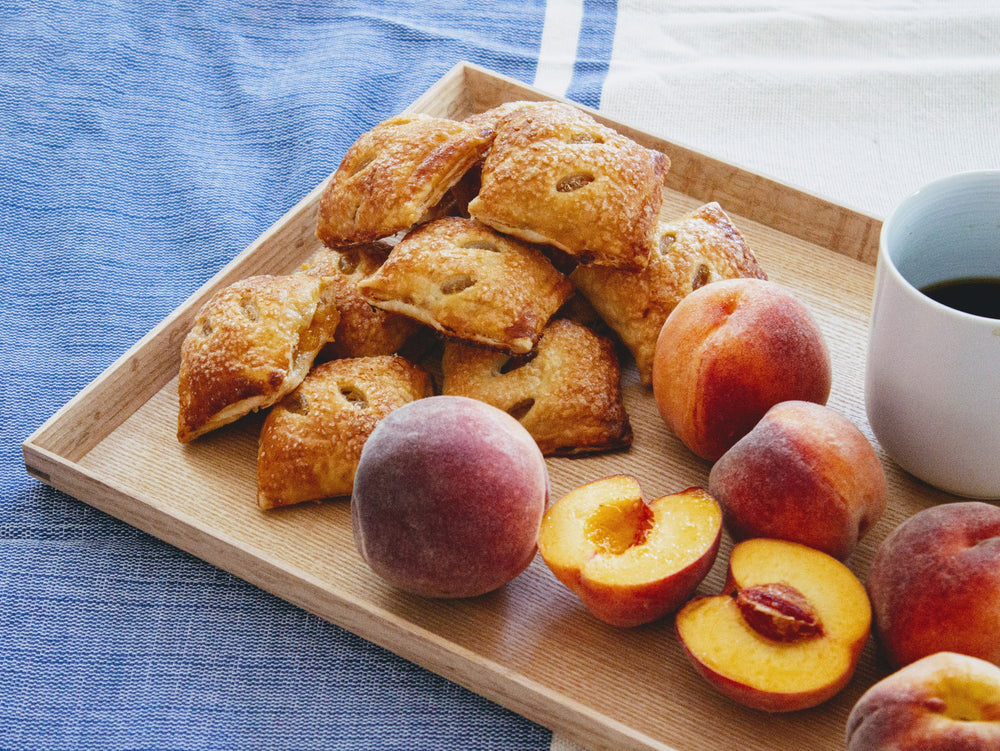 A wooden tray with peach pie bites and fresh peaches next to a cup of coffee.