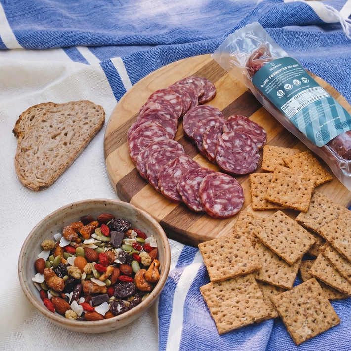 Picnic spread with sliced salami on a wooden board, square crackers, a bowl of trail mix, and bread on a blue striped cloth