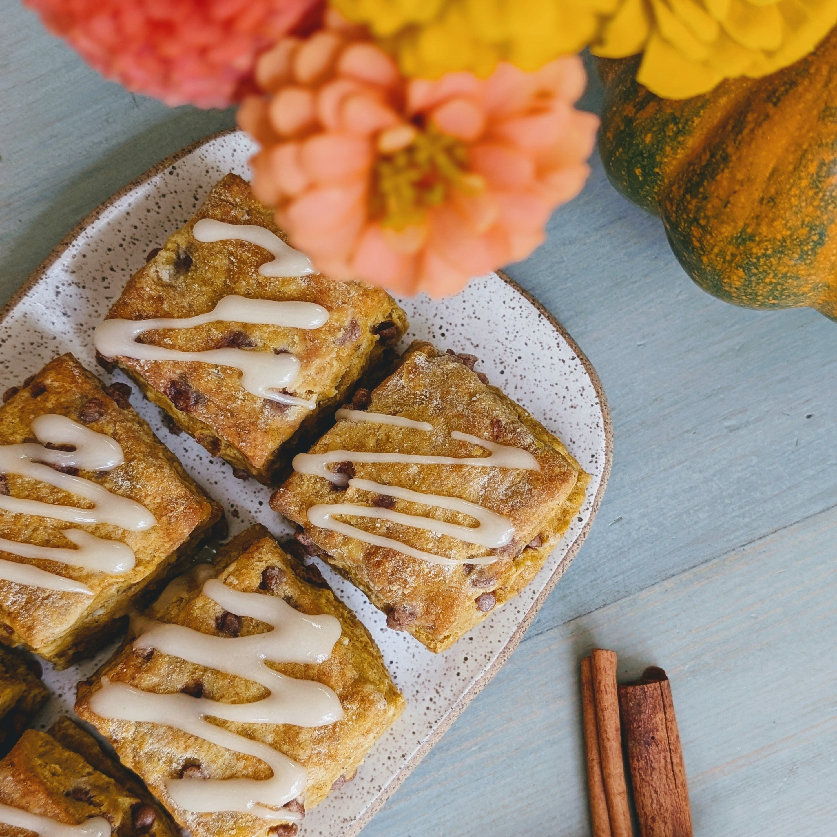 Four iced square scones on a speckled plate next to zinnia flowers, a small pumpkin, and cinnamon sticks on blue wood.
