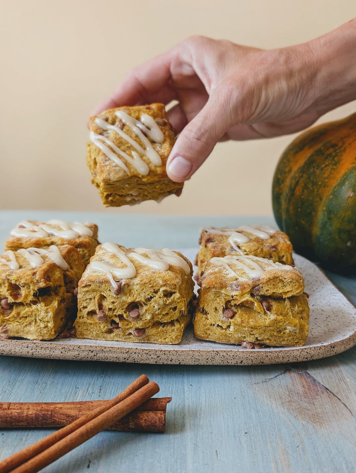 A hand reaching for a pumpkin scone drizzled with icing, surrounded by more scones and cinnamon sticks on a plate, with a decorative gourd in the background.