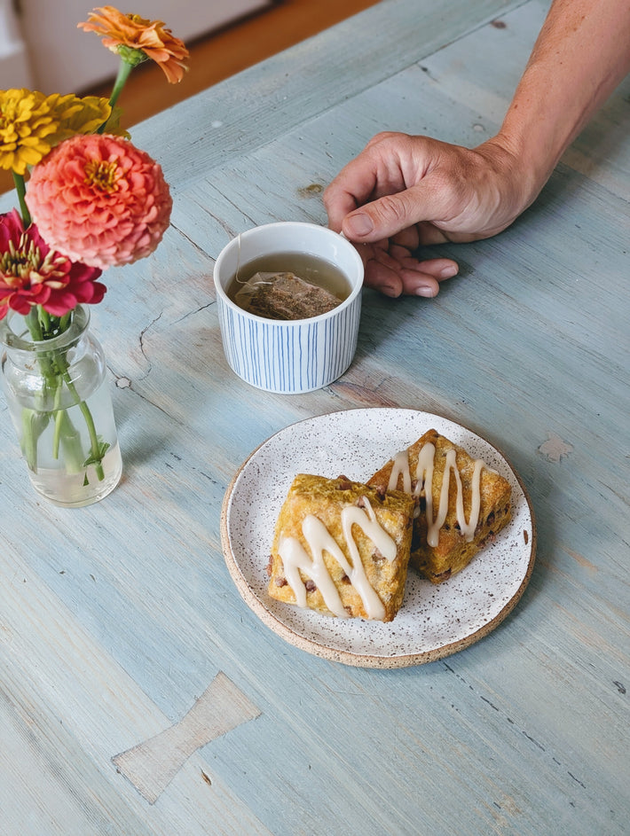A hand reaching for a cup of tea next to a plate of two iced pastries and a vase of flowers.