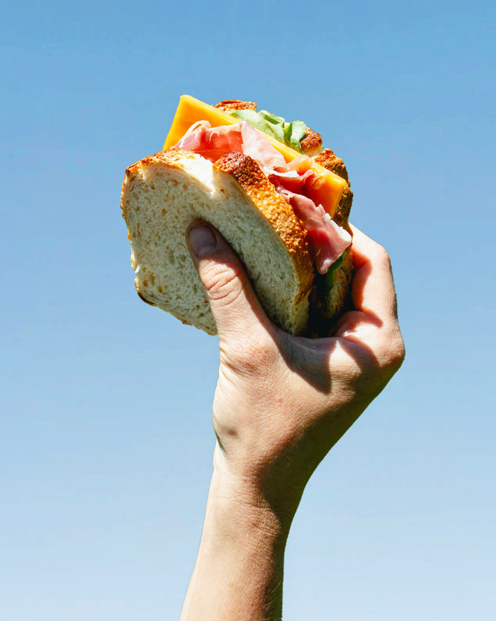 Hand holding a sandwich with layers of meat, cheese, and vegetables against a blue sky