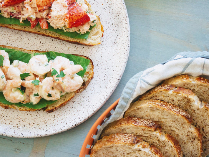 Two open-faced sandwiches with shrimp and lobster on a plate, alongside a basket of sliced bread.